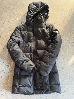 Svart lång pufferjacka från Replay - Svart pufferjacka från Replay med huva och dragkedja framtill. Jackan är lång, har två snedställda fickor med dragkedja och ribbade muddar vid ärmsluten. Klassisk quiltad look och Replay-logga på ärmen. Perfekt för kalla dagar.