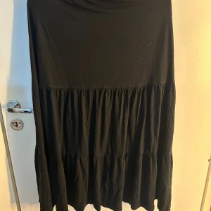 Svart långkjol i bomull - En svart långkjol med volangdetaljer och mjukt fall. Kjolen är sydd i flera sektioner för en luftig känsla och har en enkel, stilren. Obs de finns smått hål