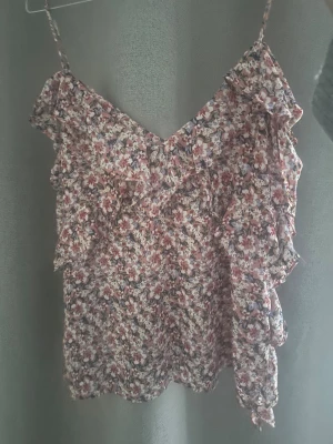 Blommig volangtop med smala axelband - Söt blommig linnetopp med volangdetaljer och smala axelband. Toppen har ett mönster i rosa, vitt och lila toner och är perfekt för varma dagar. Luftigt material och v-ringad skärning ger en romantisk vibe.