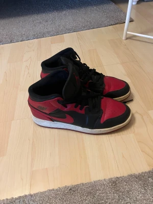 Nike Air Jordan 1 Mid Svart/Röd 39 - Säljer ett par Nike Air Jordan 1 Mid i klassisk svart och röd färg. Skorna har snörning, rund tå och vit sula. Ikonisk Jordan-logga på sidan och tungan. Tillverkade i skinn och syntetmaterial, perfekta för dig som gillar streetstyle.
