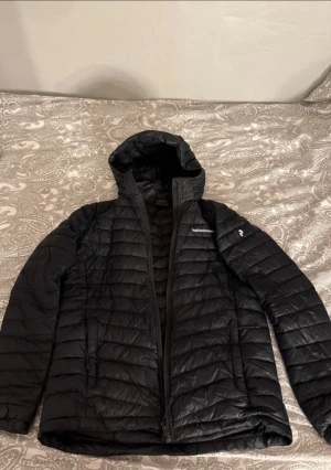 Svart dunjacka Peak Performance 170 - Svart dunjacka från Peak Performance med huva och quiltad design. Modellen heter JR Frost Down Hood Jacket, jackan är använd och har ett hål (se på bild 2) men går nog att laga 