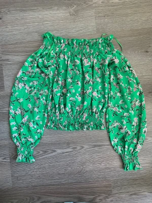 Grön blommig off-shoulder blus från Ginatricot - Säljer en grön blommig blus från Ginatricot i storlek XS. Blusen har smock upptill och nedtill, samt långa puffärmar med smock vid ärmslut. Mönstret består av små rosa och vita blommor. Perfekt för dig som gillar färgglada och romantiska plagg.