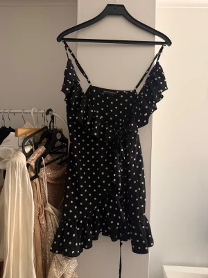 Prickig omlottklänning  - Svart kort omlottklänning med vita prickar från PrettyLittleThing. Klänningen har volangdetaljer längs urringningen och fållen, smala axelband och knyt i midjan. 