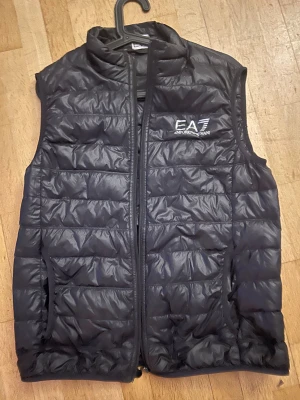 Svart dunväst från Emporio Armani EA7 - Svart quiltad dunväst från Emporio Armani EA7 med broderad logga på bröstet. Västen har ståkrage, dragkedja framtill och två sidofickor. Perfekt för lager-på-lager och snygg till streetwear. Materialet är polyester och dun.