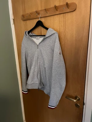 Grå Moncler zip hoodie  - Snygg grå hoodie från Moncler med huva och hel dragkedja framtill. Tröjan har Moncler-logga på ärmen och ribbade muddar med röd, vit och marinblå rand. Tillverkad i mjuk bomull, perfekt för chill dagar. Klassisk och stilren design.