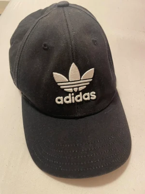 Svart Adidas keps med justerbar rem - Svart keps från Adidas med klassisk treklöver-logga broderad i vitt framtill. Kepsen har böjd skärm och justerbar rem med spänne bak för perfekt passform. Tillverkad i bomull och har Adidas-tryck på insidan. Snygg och enkel stil som funkar året runt.