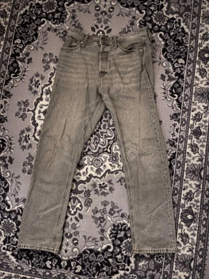 Grå jeans Relaxed Chris Jack & Jones - Säljer ett par grå jeans från Jack & Jones, modell Relaxed/Chris. De har en avslappnad passform med raka ben och klassisk femficksdesign. Jeansen är tillverkade i bomull och har en snygg tvättad look med läderpatch bak i midjan. Skick 8/10