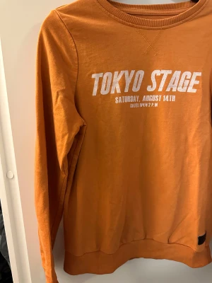 Orange sweatshirt från Lager 157 strl: 160 - Cool orange sweatshirt från Lager 157 med trycket 'TOKYO STAGE' i vitt på bröstet. Tröjan har rund hals, ribbade muddar och är tillverkad i en mjuk mix av bomull och polyester. Perfekt för dig som gillar streetstyle och snygga prints.