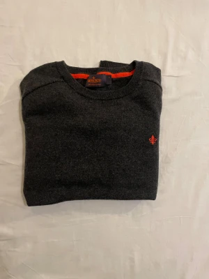 Mörkgrå merinoullströja Morris s - Snygg mörkgrå stickad tröja från Morris i 100% merinoull. Klassisk rund hals och långärmad modell med orange broderad logga på bröstet. Perfekt för dig som gillar stilrena och bekväma plagg med en liten färgdetalj.