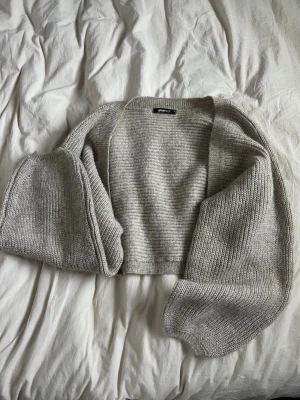 Beige stickad bolero kofta från Gina Tricot - Mysig beige stickad bolero kofta från Gina Tricot i storlek XS. Koftan har en öppen front, långärmade ärmar och en klassisk ribbad kant. Knappt använd