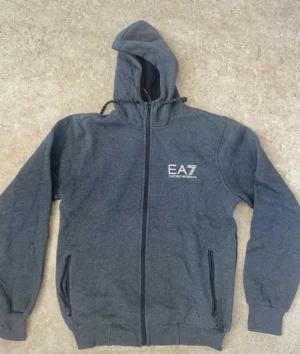 Grå hoodie EA7 Emporio Armani - Snygg mörkgrå hoodie från EA7 Emporio Armani med dragkedja framtill och två fickor med zip. Tröjan har huva med snörning och ribbade muddar vid ärmslut och nederkant. Perfekt för chill eller streetwear. Logga tryckt på bröstet.