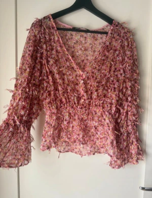 Blommig rosa blus med volangärm - Supersöt blommig blus i rosa toner med v-ringning och knappar framtill. Blusen har volangiga långa ärmar och smockad midja som ger en snygg siluett. Tunt, luftigt material – perfekt för vår och sommar. Perfekt för dig som gillar romantisk stil!