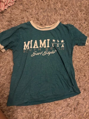 Grön t-shirt Surf Style Miami - Grön t-shirt från Surf Style med vit text 'MIAMI USA South Beach Surf Style' framtill. Blir baby tee på