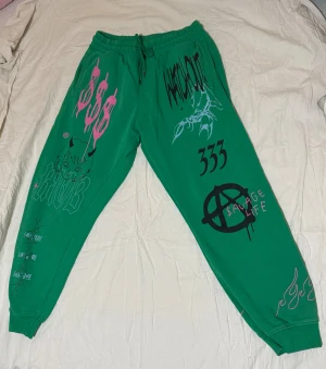 Gröna Savage Life mjukisbyxor med tryck - Säljer ett par gröna mjukisbyxor med feta grafiska tryck i rosa och svart över hela byxorna. Byxorna har snörning i midjan, muddar vid benslut och coola detaljer som eldflammor, barbed wire och olika texter. Perfekt för dig som gillar streetwear och unik stil.