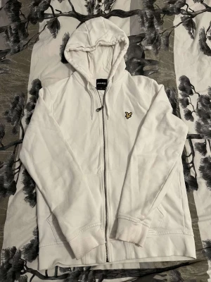 Vit hoodie från Lyle & Scott med dragkedja - Vit hoodie från Lyle & Scott med klassisk gul broderad logga på bröstet. Tröjan har huva med snörning, praktiska fickor och hel dragkedja framtill. Tillverkad i mjuk bomull och har ribbade muddar vid ärmslut och nederkant. Perfekt för chill dagar.