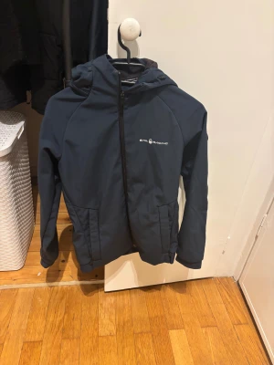 Mörkblå vindjacka från Sail Racing - Snygg mörkblå vindjacka från Sail Racing med huva och dragkedja framtill. Jackan har två fickor med dragkedja och en stilren logga på bröstet. Perfekt för blåsiga dagar och enkel att matcha med det mesta.