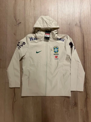 Nike Brasil vindjacka med huva - Snygg beige vindjacka från Nike med Brasilien CBF-logga och sponsormärken. Jackan har huva, hel dragkedja och meshfoder för extra komfort. Perfekt för dig som gillar fotboll och vill ha en sportig stil. Ikonisk Nike Swoosh på bröstet.