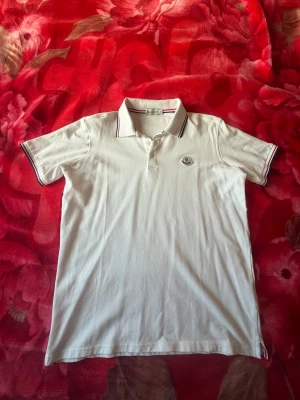 Vit Moncler piké med broderad logga - Säljer en fräsch vit pikétröja från Moncler i bomull. Klassisk krage med röd och marinblå rand, broderad Moncler-logga på bröstet och knappar framtill. Kortärmad modell med snygga detaljer på ärmslut och sidor. Perfekt för dig som gillar stilrena märkeskläder.