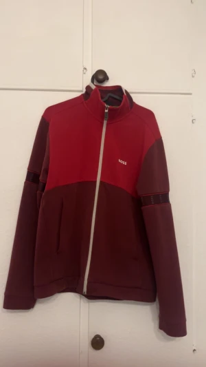 Röd Boss Dress - Snygg röd ziptröja från BOSS med kontrasterande mörkröda paneler och logoband på ärmarna. Tröjan har hel dragkedja framtill, hög krage och två fickor. Perfekt för chill eller träning, med stilren design och bekväm passform. BOSS DRESS FRÅN ROOM1006 I MALMÖ EMPORIA KÖPT FÖR 6000kr FÖRRA ÅRET LIMITED EDITONF SÄLJS EJ LÄNGRE