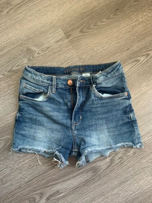 Blå high waist jeansshorts &Denim - Säljer ett par blå jeansshorts från &Denim med hög midja och råa, fransiga kanter. Klassisk femficksmodell med knapp och dragkedja framtill. Perfekta för sommarens chill dagar och har en snygg tvättad look.