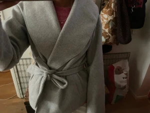 Grå omlott kappa med bälte - Säljer en stilren grå kappa med omlott-design och bälte i midjan. Jackan har bred krage och långa ärmar, perfekt för dig som gillar minimalistisk och clean look. Passar dig som vill ha en snygg och enkel jacka till hösten eller vintern.