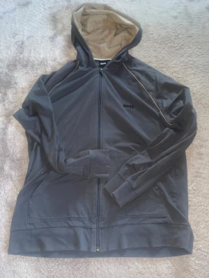 Grå hoodie från Hugo Boss, storlek L - Snygg grå hoodie från Hugo Boss i storlek L. Tröjan har huva med beige insida, dragkedja framtill och diskret BOSS-logga på bröstet. Tillverkad i mjuk bomull, perfekt för chill dagar. Långa ärmar och praktiska fickor på sidorna.