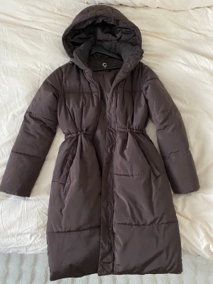 Jacka  - Säljer en brun lång pufferjacka från C i storlek XS. Jackan har en stor huva, dragkedja framtill och snyggt rynkad midja för en schysst siluett. Perfekt för kalla dagar och har två praktiska fickor på sidorna.