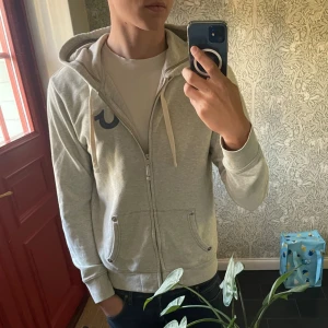 Grå hoodie från True Religion - Snygg ljusgrå hoodie från True Religion i nyskick. Kan räkna på en hand hur många gånger jag använt denna tröja, vet inte riktigt varför. Säljer nu bara för jag känner att det inte riktigt passar min garderob längre😄