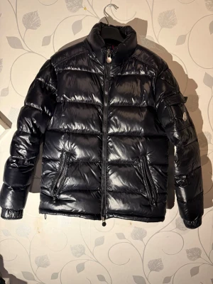 Svart Moncler pufferjacka herr - Svart pufferjacka från Moncler med glansig finish och klassisk logga på ärmen. Jackan har hög krage, två framfickor med dragkedja och en ficka på ärmen. Perfekt för kalla dagar och har en riktigt schysst streetstil. Köpt på plick för cirka 2 år sedan och då var den helt ny, säljer då den är för liten för mig. Är väldigt säker på att den är äkta men kan inte vara hundra eftersom den köptes på plick. Skriv vid frågor funderingar mm mvh🙌