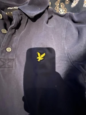 Marinblå pikétröja från Lyle & Scott - Snygg marinblå pikétröja från Lyle & Scott med klassisk krage och tre knappar framtill. Tröjan har det ikoniska gula fågelbroderiet på bröstet och är gjord i mjuk bomull. Perfekt för en chill och stilren look.
