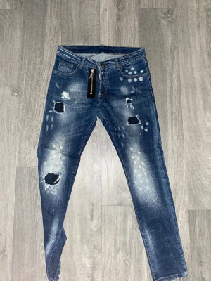 Blåa Dsquared jeans - Säljer ett par blå jeans från Dsquareds med coola slitningar, hål och vita prickar på framsidan. Jeansen har rak passform, klassiska fem fickor och ett tryck med texten 'BE COOL BE NICE' på bakfickan. Perfekt för dig som gillar streetstyle.