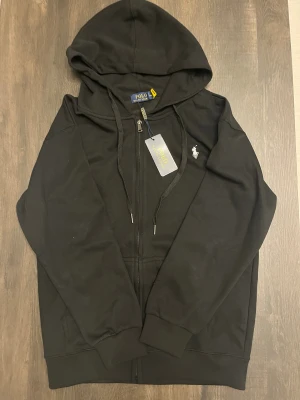 Svart hoodie från Polo Ralph Lauren - Svart hoodie från Polo Ralph Lauren med klassisk vit broderad logga på bröstet. Tröjan har huva med snörning, dragkedja framtill och ribbade muddar. Tillverkad i mjuk bomull för skön komfort. Perfekt för en chill och stilren look.