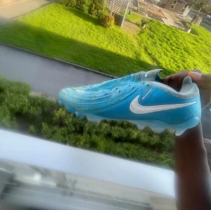 Blå Nike fotbollsskor med vit swoosh - Säljer ett par blå Nike fotbollsskor med vit swoosh på sidan som är elites med  coola detaljer. Skorna har snörning och är designade för att ge bra grepp på planen. Ovandelen är i syntetmaterial och sulan har dobbar för bästa fäste. Perfekta för dig som vill sticka ut på fotbollsplanen. Boxen och gympa påsen ingår med köpet 