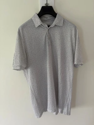 Gråmönstrad golfpiké från Galvin Green - Snygg grå/vit golfpiké från Galvin Green med diskret mönster över hela tyget. Klassisk krage, kort ärm och knappar framtill. Perfekt för golfbanan eller chill dagar. Liten logga i nacken och på ärmen. Tillverkad i mjukt och svalt material. Storlek: L passar M. Nypris: 999kr