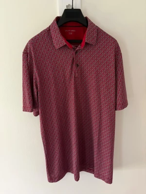 Röd mönstrad golfpiké från Galvin Green - Säljer en röd golfpiké från Galvin Green med små, täta mönster i vitt och svart. Skjortan har klassisk krage, korta ärmar och knappar framtill. Perfekt för dig som gillar sportig och stilren look. Perfekt till golfbanan. Storlek: L passar M. Nypris: 999kr