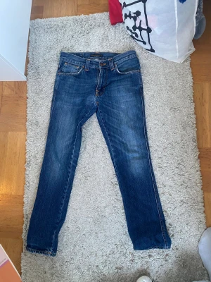 Blå Nudie Jeans, W29 L30 - Säljer ett par klassiska blå jeans från Nudie Jeans med snygga kontrastsömmar och ikonisk broderad detalj på bakfickorna. Modellen har normal passform och raka ben, tillverkade i slitstarkt bomullsjeans. Perfekta för dig som gillar stilrena och tidlösa jeans.
