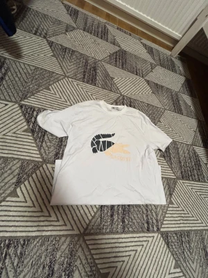 Vit Lacoste t-shirt med grafiskt tryck - Vit t-shirt från Lacoste i bomull med ett stort grafiskt krokodiltryck i svart och beige på bröstet. Klassisk rund halsringning och korta ärmar. Perfekt för dig som gillar stilrena plagg med en twist.