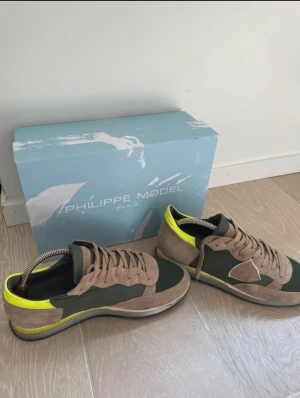 Philippe Model sneakers brun/grön - Snygga sneakers från Philippe Model i brun mocka och grönt tyg med detaljer i neon-gult skinn. Skorna har snörning, rund tå och platt sula. Stilren design med kontrasterande färger och logotyp på sidan. Perfekt för dig som gillar unika och trendiga sneakers. Priset sitter inte fast i sten.