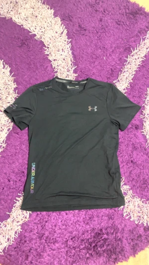 Svart Under Armour HeatGear t-shirt - Svart t-shirt från Under Armour med reflekterande logga och textdetaljer i regnbågsfärger. Modellen är kortärmad och har en rund hals. Materialet är lätt och andas, perfekt för träning och löpning. Snygg slim passform som sitter skönt mot kroppen.
