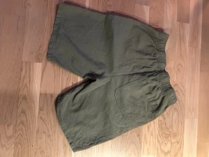 Olivgröna loose linneshorts - Säljer ett par olivgröna linneshorts i loose fit med elastisk midja och knapp framtill. Shortsen har två sidofickor och en bakficka, tillverkade i svalt linnematerial. Perfekta för varma dagar när du vill ha en chill och avslappnad stil.