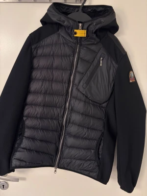 Svart pufferjacka från Parajumpers - Snygg svart pufferjacka från Parajumpers med huva, gul detalj vid halsen och patch på ärmen. Jackan har quiltad framsida och rygg, släta ärmar och flera dragkedjeförsedda fickor. Perfekt för dig som vill ha en sportig och stilren look.