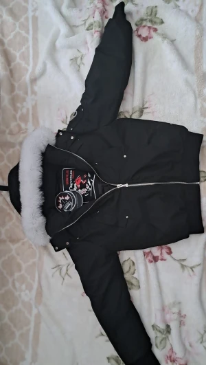 Svart dunjacka Moose Knuckles M - Säljer en svart dunjacka från Moose Knuckles med fluffig vitt fuskpäls runt huvan. Jackan har dragkedja, flera fickor och coola detaljer på insidan. Perfekt för kalla dagar och riktigt snygg streetstil.Har scan och är helt ny med etikett på,äkta men det finns inget som bevisar att den är äkta 