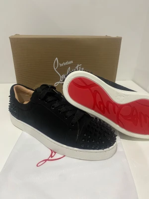 Christian Louboutin svarta sneakers med nitar - Svarta sneakers från Christian Louboutin med mattsvarta nitar på tå och häl, klassisk röd sula och svarta skosnören. Ovandel i mocka och innerfoder i beige skinn. Snygg och edgy design som sticker ut med exklusiva detaljer.