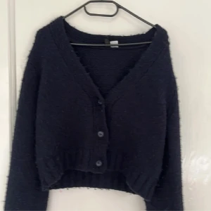 Mörkblå kofta med knappar - Mysig mörkblå croppad kofta med v-ringning och tre knappar framtill. Koftan har långärmade ärmar och ribbad kant vid midjan och ärmslut. Perfekt för lager-på-lager och chill dagar. Materialet är stickat och ger en skön känsla.