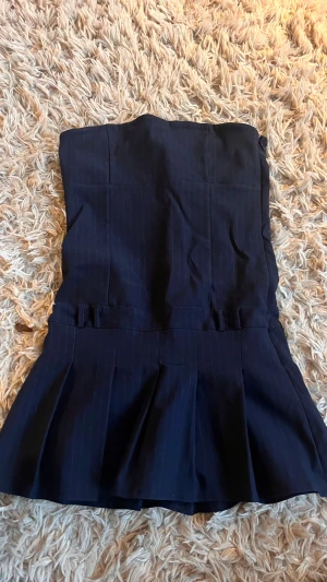 Mörkblå randig känning  från Boohoo - Snygg mörkblå klänning från Boohoo med diskreta ränder och låg midja. Modellen har en kjolliknande design med breda veck framtill som ger en cool och unik look. Perfekta för dig som gillar att sticka ut med stilrena detaljer. Även shorts under