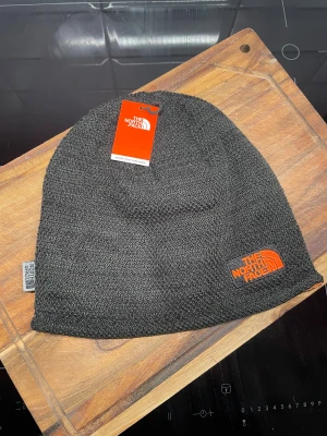 Svart mössa från The North Face - Säljer en svart mössa från The North Face med orange broderad logga framtill. Utsidan är stickad och insidan är fodrad med mjuk orange fleece för extra värme. Perfekt för kalla dagar och snygg streetstil. Oanvänd 