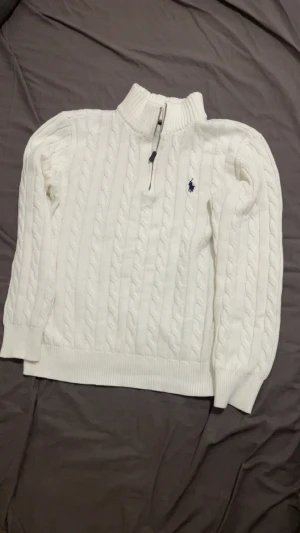 Vit stickad tröja från Ralph Lauren - Säljer en vit stickad tröja från Ralph Lauren med half zip och kabelstickat mönster. Tröjan har långärmad design, ribbade muddar och en liten blå logga broderad på bröstet. Perfekt för dig som gillar klassisk och clean stil.