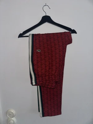 Röda mönstrade mjukisbyxor Lacoste M - Snygga röda mjukisbyxor från Lacoste i regular fit med geometriskt mönster och klassisk krokodillogga. Byxorna har snörning i midjan och vita/gröna ränder längs sidorna. Perfekt för chill eller streetwear.