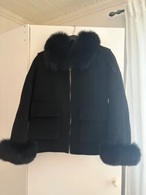 Svart pälsjacka med pälskrage - Snygg svart pälsjacka med fluffig pälsdetalj på kragen och ärmsluten. Jackan har två stora fickor framtill och stängs med dragkedja. Modellen är loose fit och har en stilren look som sticker ut med sina pälsdetaljer. Materialet är kashmir och pälsen är rävpäls. Köpt från Garoff för omkring 3500