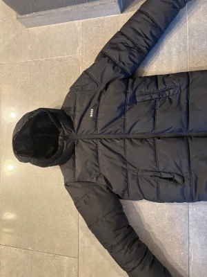 Svart pufferjacka från BOSS med huva - Säljer en svart pufferjacka från BOSS med huva och dragkedja. Jackan har två sidofickor med dragkedja och diskreta BOSS-loggor på bröstet och ärmen. Perfekt för kalla dagar och har en stilren, modern look.